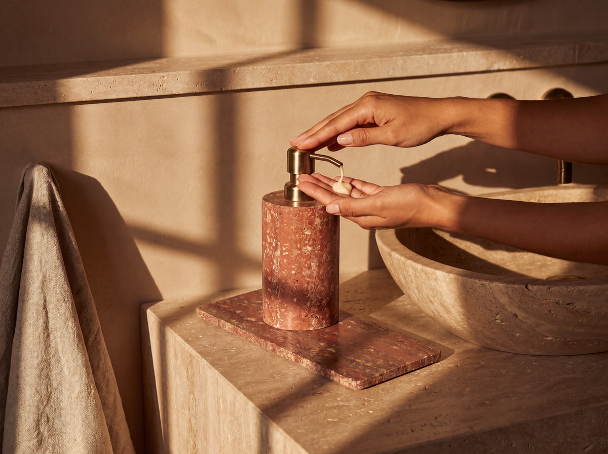 Woman pressing the brass pump of a red travertine dispenser at a stone bathroom vanity, dispensing lotion into her palm