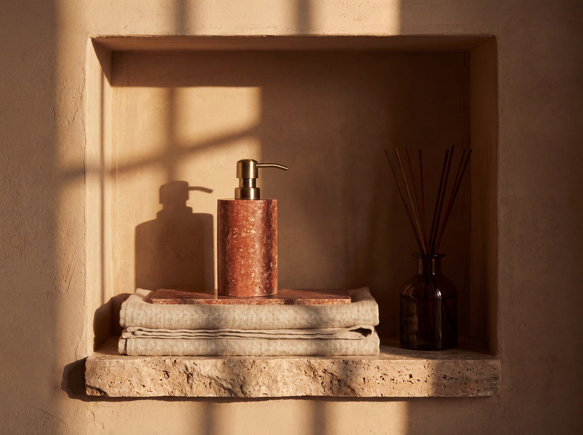 Red travertine dispenser and tray in a plaster wall niche above folded linen towels with a reed diffuser