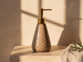 Cone-shaped ceramic soap dispenser with cocoa-brown spotted glaze and gold pump on a travertine ledge in golden window light.
