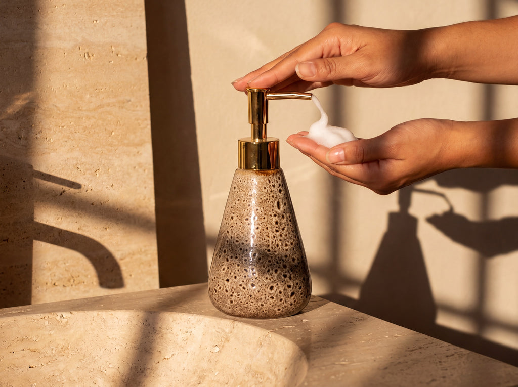 A hand pressing the gold pump of the 16 cm ceramic soap dispenser above a stone sink.